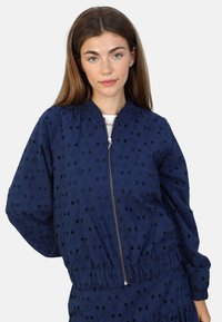 Marineblå bomberjakke med lynlåsfortil, prydet med en broderet tekstur med udskårne blomsterdesigns samt elastiske manchetter og talje.