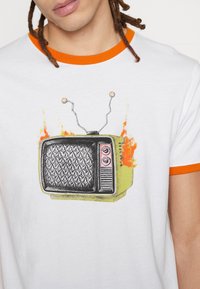 Mand iført en hvid T-shirt med orange kant, der viser et retro tv-graphic, der udsender statisk støj og flammer på begge sider.