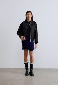 Μαύρο μπουφάν τύπου bomber, εφαρμοστή μαύρη τοπ με κοψίματα, υφασμάτινη μίνι φούστα σε σκούρο μπλε με λευκή επένδυση και μαύρες μπότες μέχρι το γόνατο με λευκές κάλτσες.