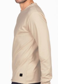 Beige Langarmhemd aus glattem Stoff. Mit Rundhalsausschnitt, schlanker Passform und einem kleinen schwarzen Etikett an der unteren Seitennaht.