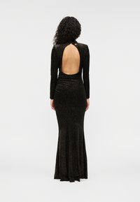 Μαύρο φόρεμα με μακριά μανίκια και εφαρμοστή γραμμή, με άνοιγμα στην πλάτη και επεκτεινόμενο κάτω μέρος, κατασκευασμένο από υφή υφάσματος με γυαλιστερή επιφάνεια.