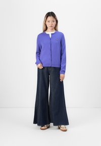 Cardigan violet avec encolure ronde, fermeture à boutons et un petit logo. Associé à un jean en denim foncé à jambes larges et des chaussures légères.