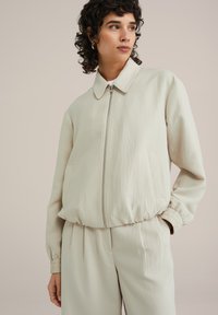 Šviesiai beige bomber tipo striukė su apykakle, užtrauktuku, elastingomis rankovėmis ir šoninėmis kišenėmis, pagaminta iš glotnaus, lengvo audinio.