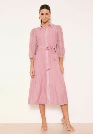 Nő rózsaszín, mintás pöttyös midi ruhában, háromnegyedes ujjakkal, gombos elöl, megkötős derékrésszel, és bézs magas sarkú szandálban, állva.