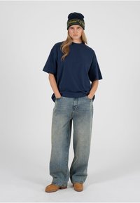 Camiseta de algodón azul marino, de manga corta, combinada con vaqueros anchos de mezclilla azul claro y zapatos marrones sin cordones. Gorro negro con detalle de texto en verde.