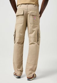 Kaki cargobyxor med flera bakfickor, rakt ben-design, slitstarkt tyg och synlig varumärkeslapp. Fötter i bruna och gula sneakers.