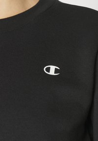 T-shirt nera in cotone con collo rotondo, caratterizzata da un piccolo logo circolare bianco sul lato sinistro del petto. Texture liscia.
