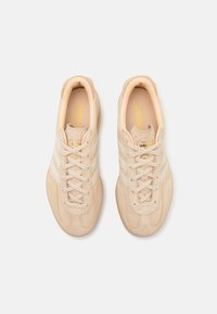 Ténis de camurça bege com três riscas brancas, biqueira redonda e sola texturizada. Apresentam atacadores bege e marca dourada na língua.