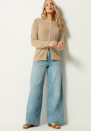 Nainen, joka pukeutuu beigeen nepparilliseen neuletakkiin, leveälahkeisiin vaaleansinisiin farkkuihin ja ruskeisiin sandaaleihin, seisoo vaaleanväristä seinää vasten.