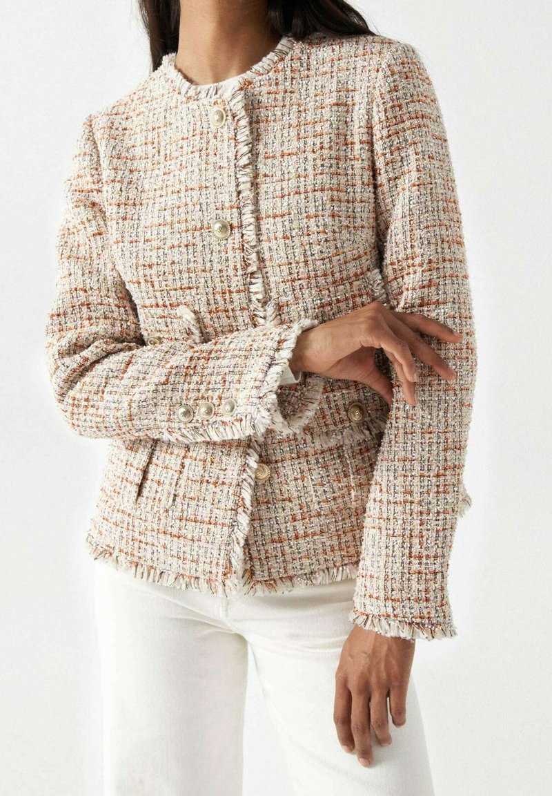 Femme portant une veste en tweed à carreaux crème et orange avec des bords effilochés, des boutons nacrés et un pantalon blanc.