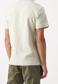 Beige polo triko s krátkým rukávem a texturovaným, ribbovaným povrchem, s plochým límečkem a rovnými lemy, kombinované s olivově zelenými cargo šortkami.