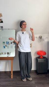 Model îmbrăcat într-un tricou alb din bumbac și pantaloni largi gri stă lângă o oglindă, alături de o lampă roșie și o valiză gri.