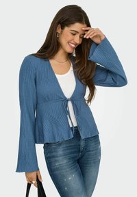 Blaues geripptes Cardigan mit langen Glockenärmeln und einem Bindeband in der Taille, über einem weißen Oberteil getragen und mit Jeans kombiniert.