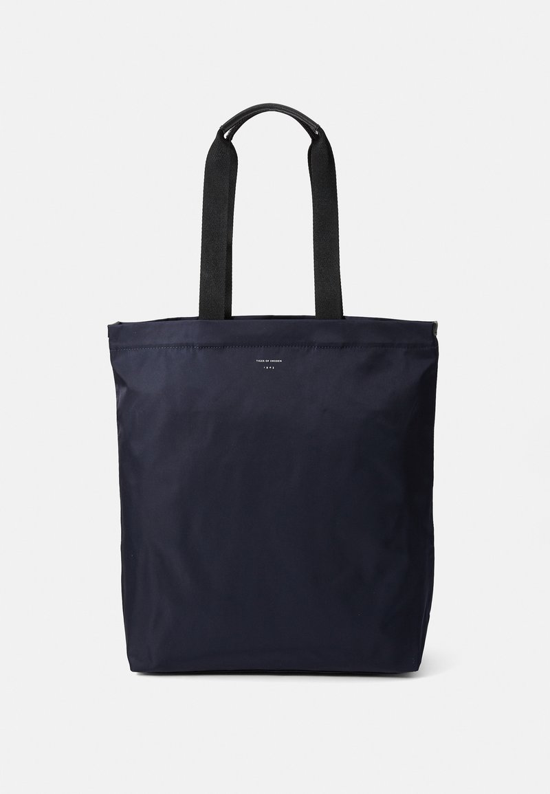 Sacola de ombro em nylon azul-marinho com alças pretas em fita, formato retangular, design minimalista, apresentando um pequeno logotipo e textura suave.