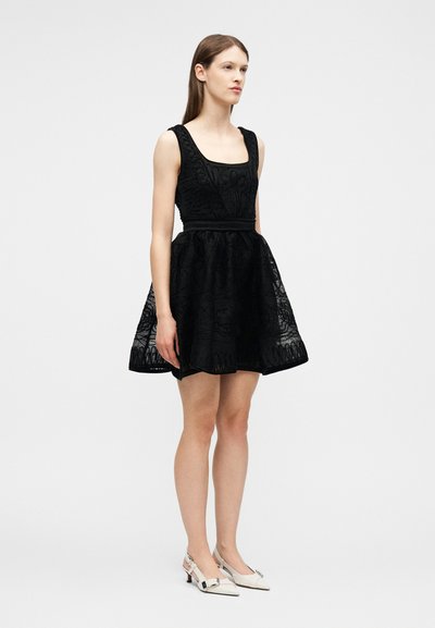 Γυναίκα στέκεται φορώντας μαύρο δαντελένιο φόρεμα χωρίς μανίκια με φουσκωτή φούστα και λευκά χαμηλά παπούτσια με μυτερή μύτη, μπροστά σε απλό φόντο.