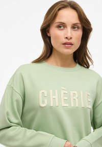 Felpa verde chiaro con la scritta "CHÈRIE" in lettere rialzate color crema. Presenta un colletto rotondo e una vestibilità rilassata. Tessuto morbido, maniche lunghe.
