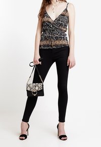 Donna con top avvolgente a motivo serpente, pantaloni skinny neri, sandali con tacchi alti neri, che tiene in mano una borsa nera con motivo serpente.
