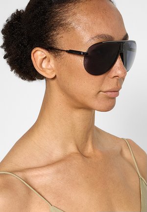 Donna con capelli ricci indossa occhiali da sole grandi e scuri e un top beige con spalline sottili, guarda verso destra su uno sfondo semplice.