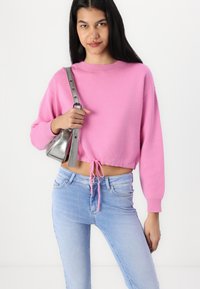 Sweater rose cropped avec un col rond et un ourlet à cordon de serrage, associé à un jean bleu clair. Le modèle tient un sac à main en argent avec une bandoulière longue.