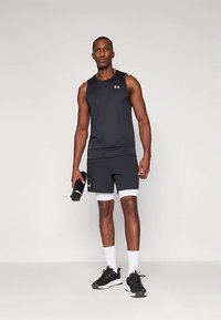 Atletický muž v čiernom tielku bez rukávov a krátkych nohaviciach, biele kompresné šortky, držiaci čiernu fľašu na vodu, stojaci na jednofarebnom pozadí.