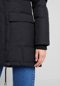 Chaqueta aislante negra con un diseño cosido, puños elásticos, grandes bolsillos y un dobladillo con cordón. La tela parece suave y duradera.