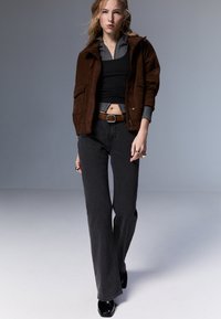 Ruskea mokkajakku, jossa on huppu, kerrosmustan toppauksen ja harmaan paidan päällä. Mustat levenevät farkut ja kiiltävät mustat saappaat viimeistelevät asun.