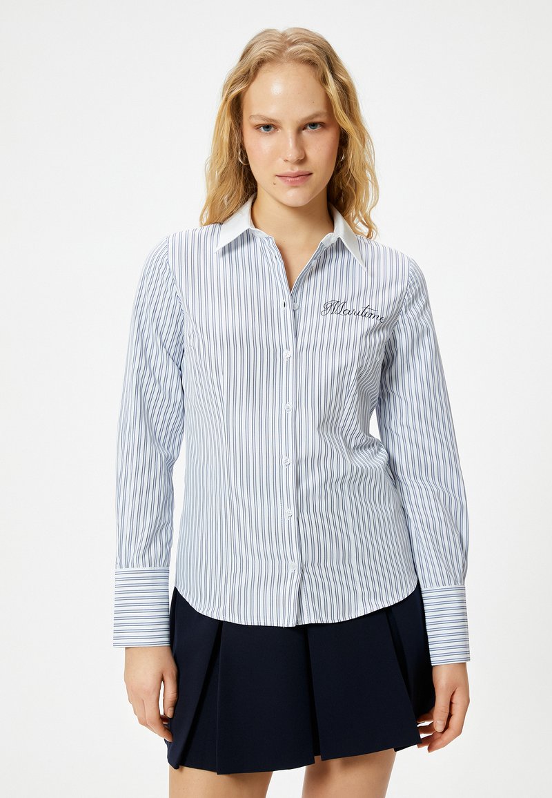 Camicia a righe blu e bianche con colletto a punta e testo ricamato. Maniche lunghe con polsini a righe contrastanti.