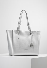 Srebrna torba na ramię wykonana z teksturowanego materiału, z podwójnymi uchwytami, zewnętrzną kieszenią i okrągłym charms'em z logo.
