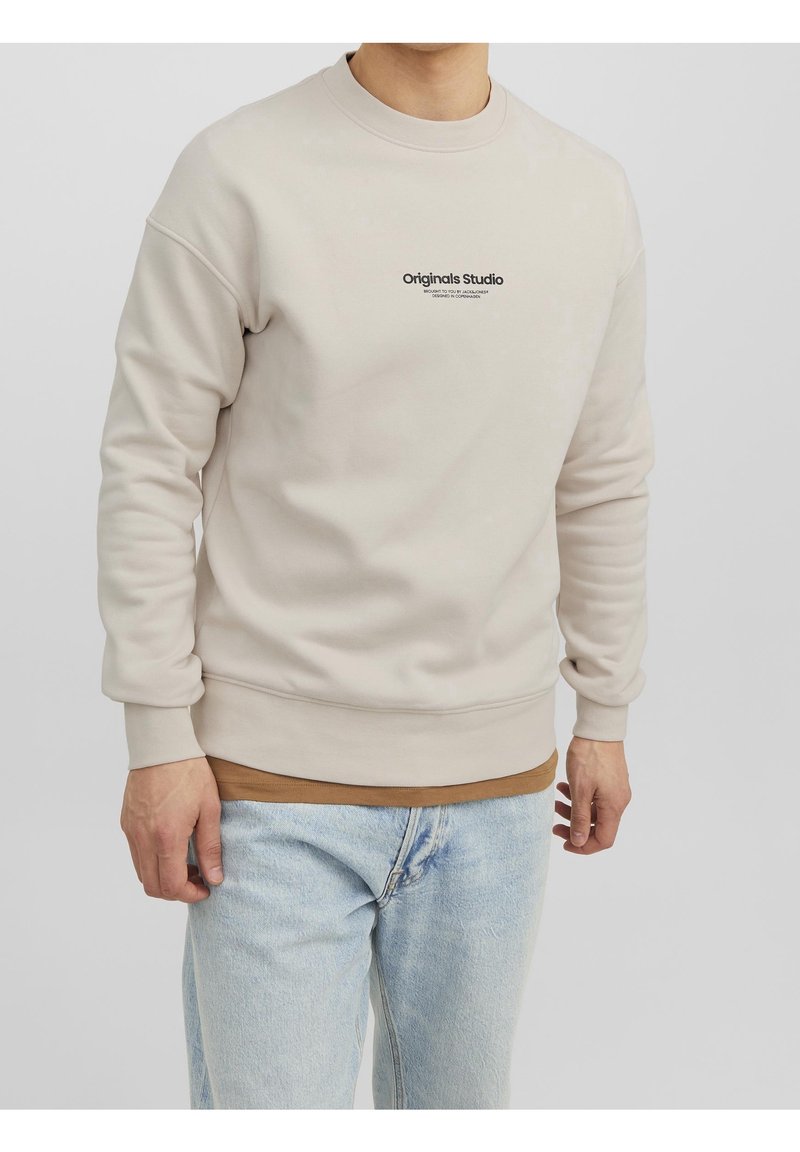 Homme portant un sweat-shirt beige avec le texte "Originals Studio" sur un sous-pull marron et un jean bleu clair, debout devant un fond uni.