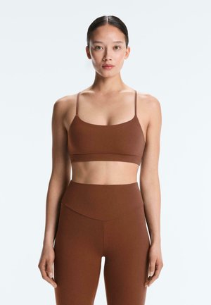 Kvinde med mørkt hår sat op i en knold, iført en brun sports-bh og leggings med høj talje, stående mod en ensfarvet lys baggrund.