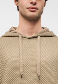 Μπεζ καπιτονέ φούτερ με κουκούλα και κορδόνια, υφασμάτινη υφή και ανοιχτόχρωμα κορδόνια. Κοντινή λήψη που εστιάζει στη λαιμουδιά και τις λεπτομέρειες της κουκούλας.