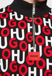 Top negro con cuello alto que presenta un patrón de letras rojas y blancas que deletrean "HUGO" y una etiqueta de logo roja prominente en la parte delantera. Cierre con cremallera visible.