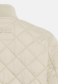 Gepolsterte beiger Jacke mit einem gerippten Kragen und gestepptem Rautenmuster. Weiche Textur mit einem minimalistischen Design und subtilen Branding-Element.