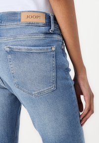 Svijetloplave traperice viskog struka, s tamnim kožnim etiketu s "JOOP!" logotipom i setom stražnjih džepova. Glatka tekstura.