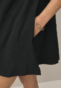 Zwarte, textuurstof jurk met een subtiel gestreept patroon, voorzien van een zijzak en een hand versierd met een zilveren kettingarmband.