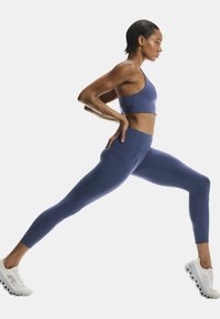 Kvinde i blå sportsbeklædning og hvide sneakers, der strækker sig i en bred lungestilling med hænderne på hofterne foran en ensfarvet hvid baggrund.