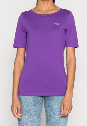 Osoba w prostym fioletowym t-shircie z krótkim rękawem, z małym białym napisem na piersi oraz jasnoniebieskich dżinsach typu acid-wash.