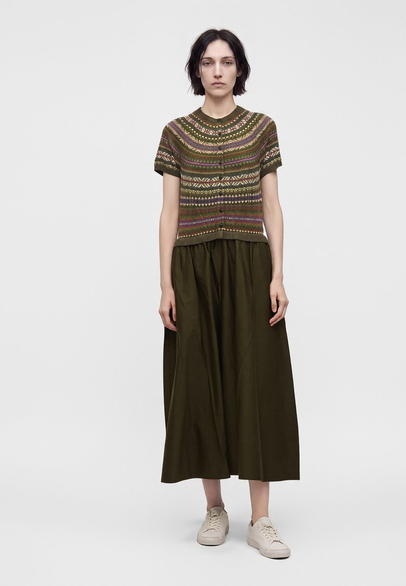 Femme debout portant un pull à manches courtes vert à motifs, une longue jupe vert foncé et des baskets beiges sur un fond clair uni.