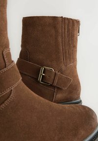 Ingrandimento di stivali alla caviglia in suede marrone con fibbia laterale e dettagli di cucitura su sfondo chiaro.