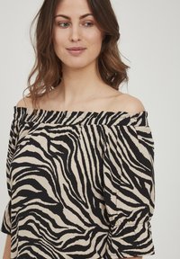 Blusa off-the-shoulder in beige e stampa zebra nera, con scollatura arricciata e maniche a sbuffo con polsini elastici. Texture del tessuto morbida.