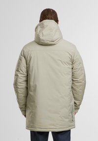 Beige isolerad jacka med huva, har en slät yta, justerbara ärmslut och en sidodragkedjeficka på nedre vänster sida.