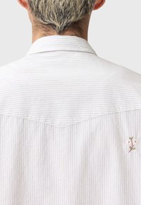 Λευκό πουκάμισο με διακριτικές κάθετες ρίγες· διαθέτει μυτερό γιακά και κεντημένο λουλουδάτο μοτίβο στην πλάτη κοντά στη γραμμή του λαιμού.