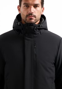 Man in een zwarte winterjas met capuchon, voorzien van een ritszak op de borst en een knoopsluiting aan de voorkant, die recht vooruit kijkt.