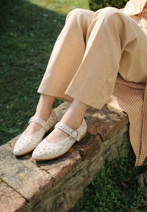 Persona con scarpe basse Mary Jane beige con borchie e pantaloni beige chiaro seduta su un bordo di mattoni ricoperto di muschio all'aperto.