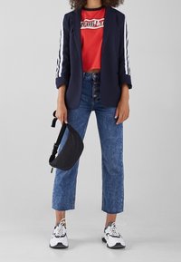 Marinblazer med vita ränder, röd grafisk crop top, högmidjade blå jeans med knappar, svart midjeväska och vita sneakers med svarta accenter.
