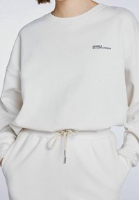 Sudadera blanca con cuello redondo acanalado y corte oversize. Presenta una pequeña impresión negra "DISEÑADO EN" en el pecho y una cinturilla elástica con cordón.
