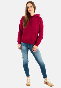 Sudadera con capucha color granate con cordón y bolsillo delantero, combinada con vaqueros azul claro y zapatillas beige con detalles granate en los laterales.