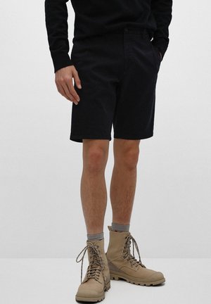 Person iført sorte shorts, sort langærmet top, grå sokker og beige snørestøvler, der står mod en ensfarvet baggrund.
