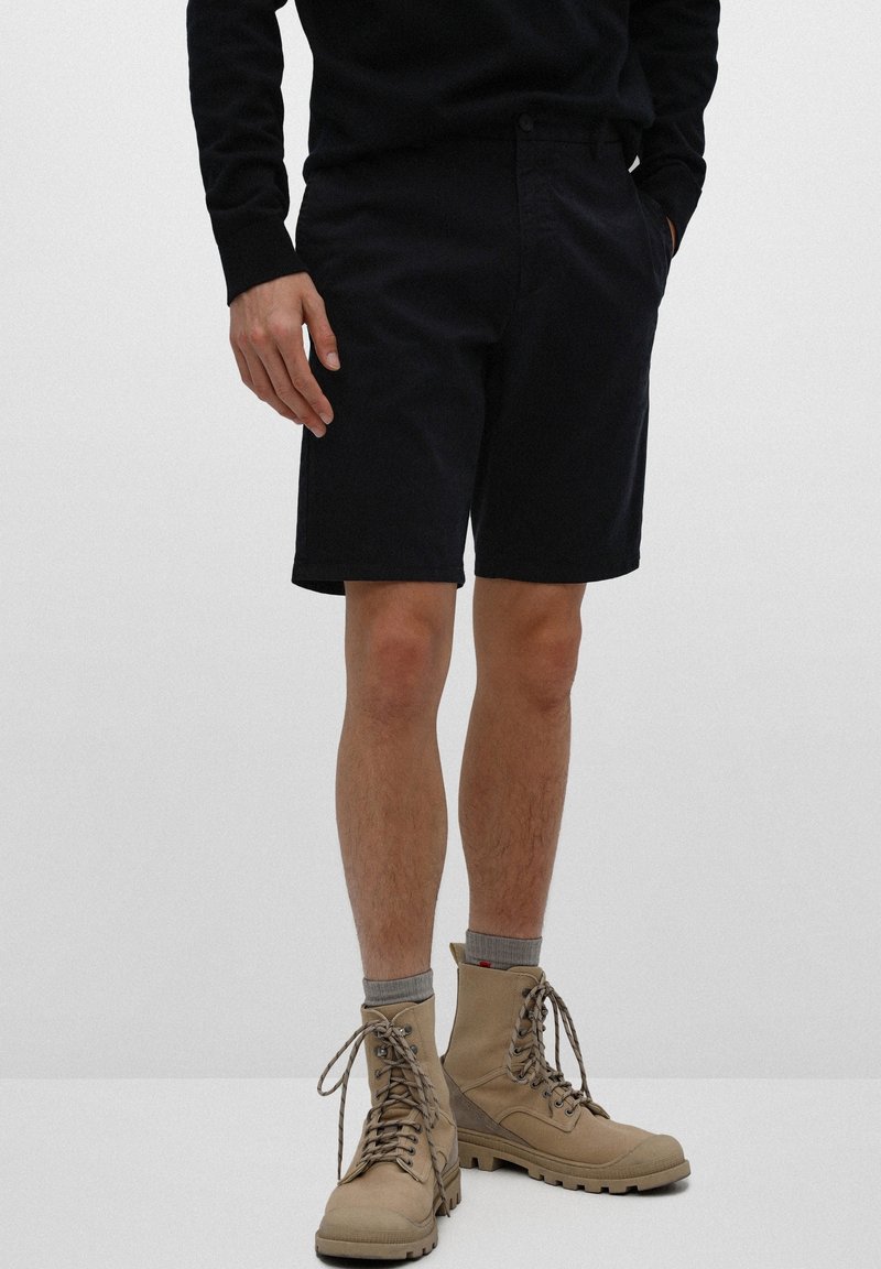 Personne portant un short noir, un haut à manches longues noir, des chaussettes grises et des bottes beiges à lacets, debout devant un fond uni.