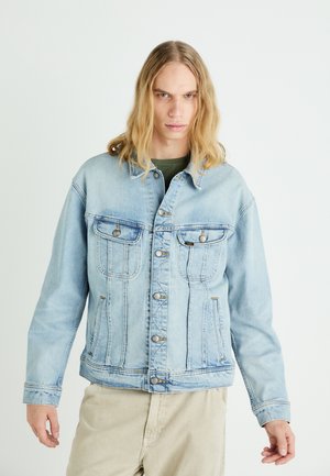 Młoda osoba z długimi blond włosami, ubrana w jasnoniebieską jeansową kurtkę i beżowe spodnie, stoi na jednolitym jasnym tle.